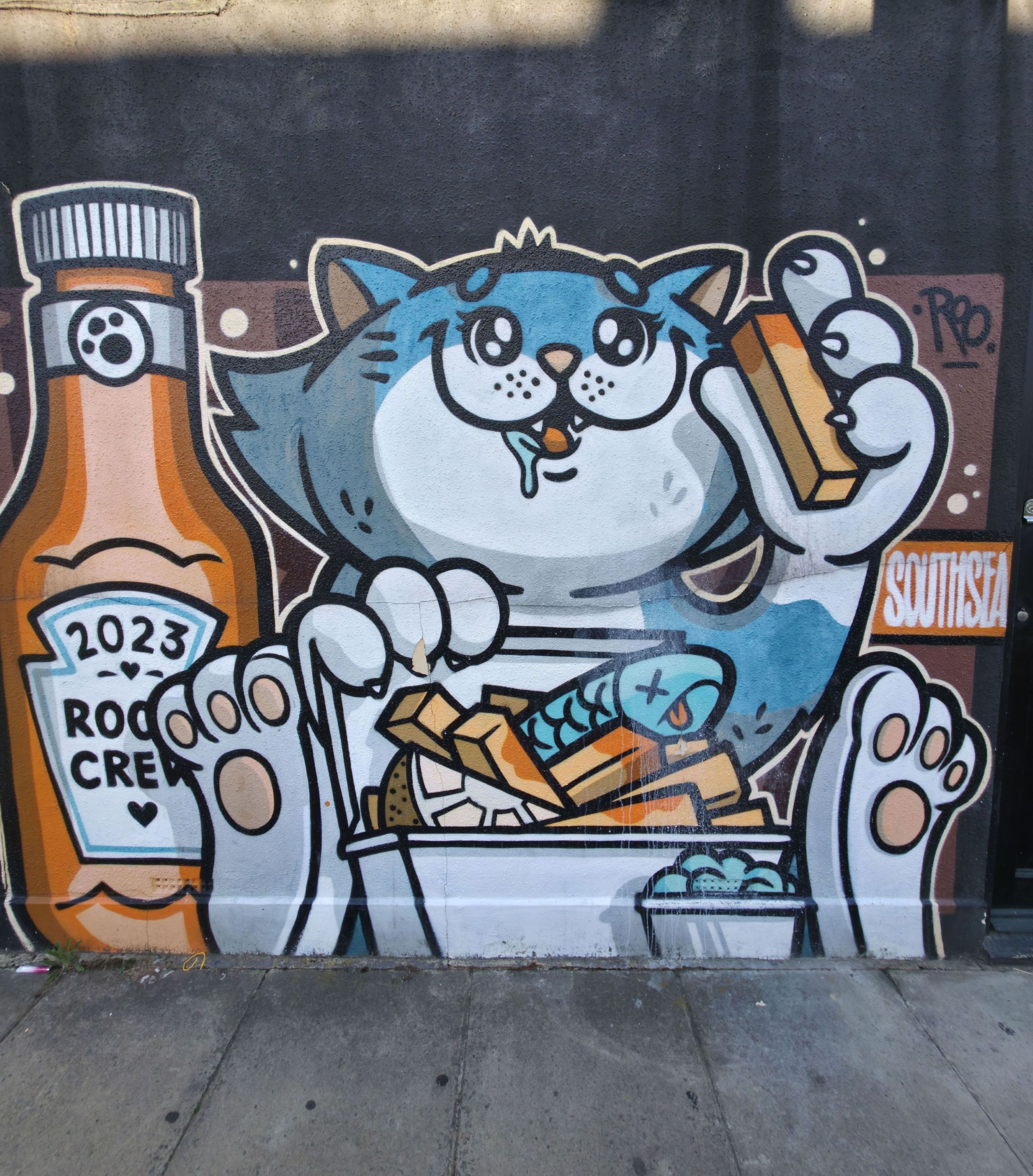 Photo of a cat eating fish and chips with a bottle of sauce, found in Albert Road, Southsea, Hampshire, United Kingdom. Photo taken by Baldi, A.K.A Amonn AL-Mahrouq.